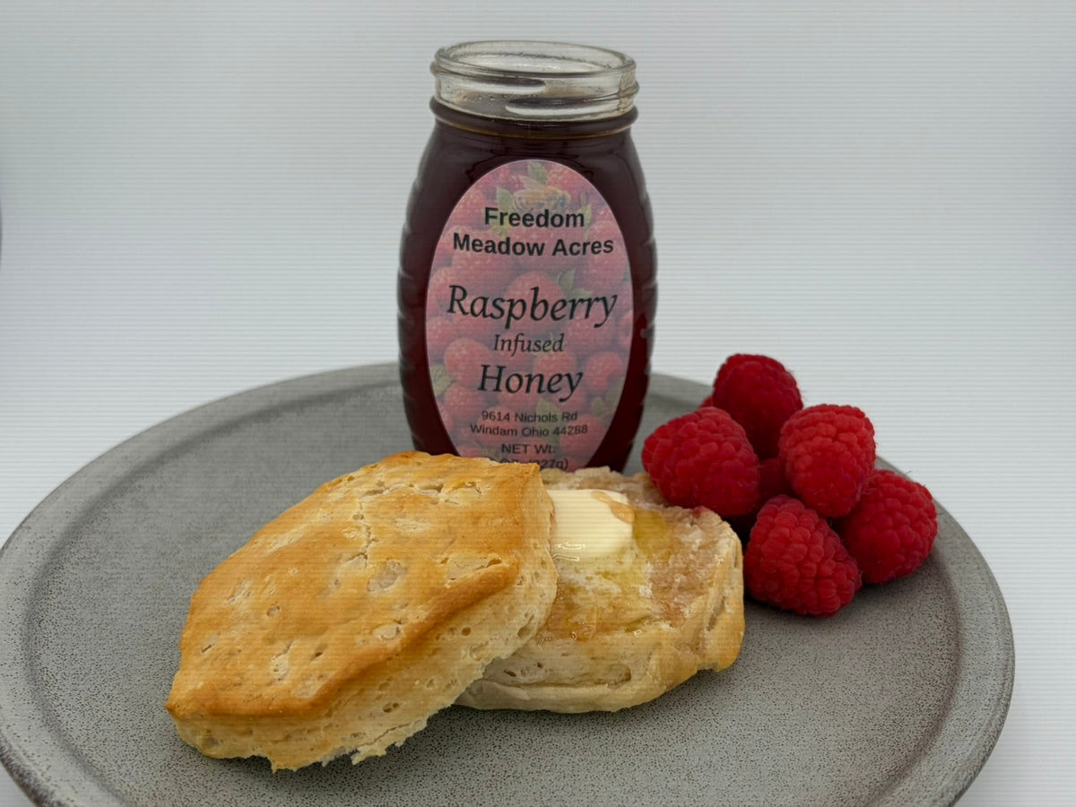 A glass jar of Freedom Meadow Acres Raspberry Infused Honey stands behind a gray plate holding two golden-brown biscuits with butter and fresh raspberries.