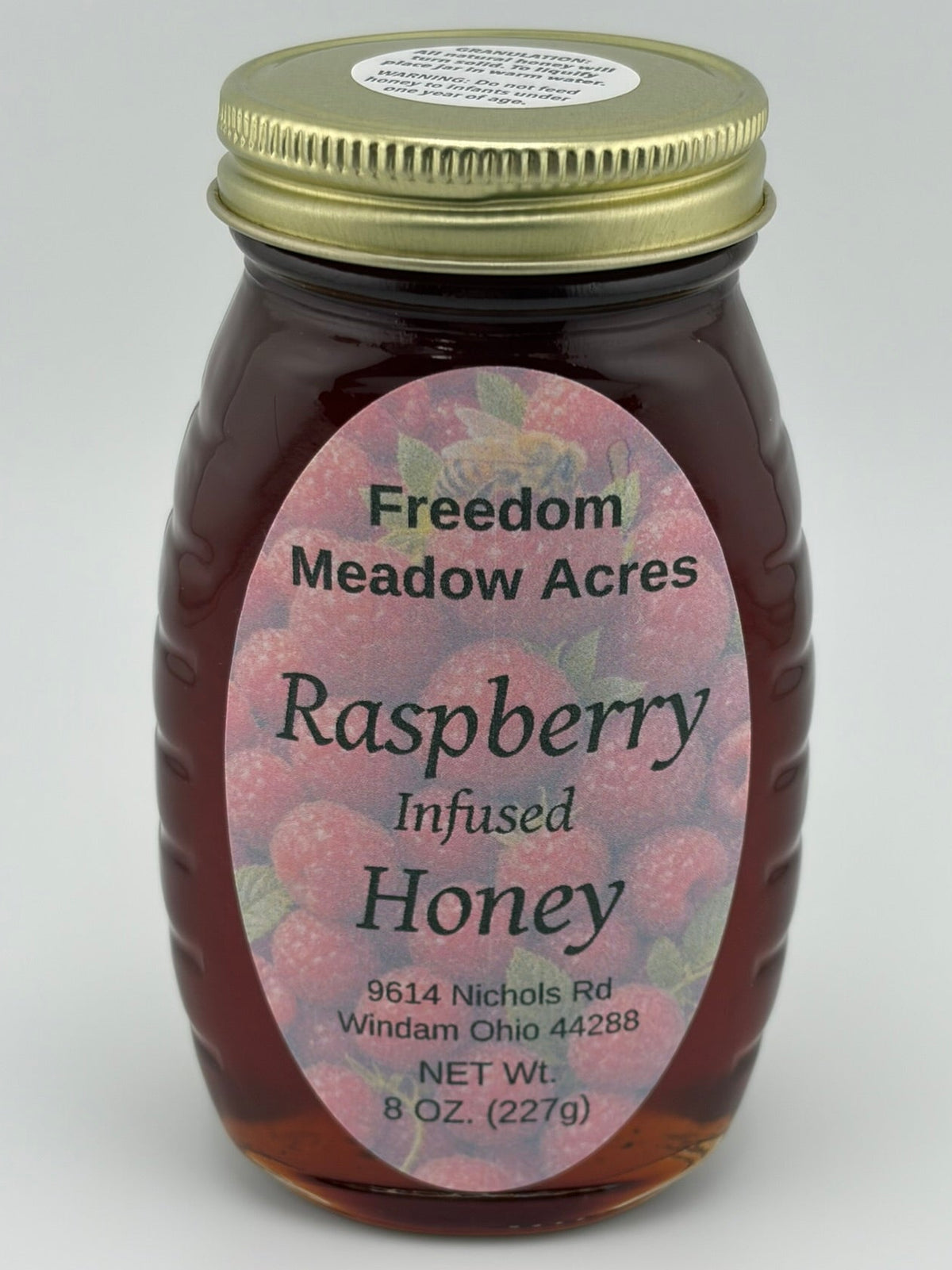 An 8 oz glass jar of Freedom Meadow Acres Raspberry Infused Honey with a gold metal lid.