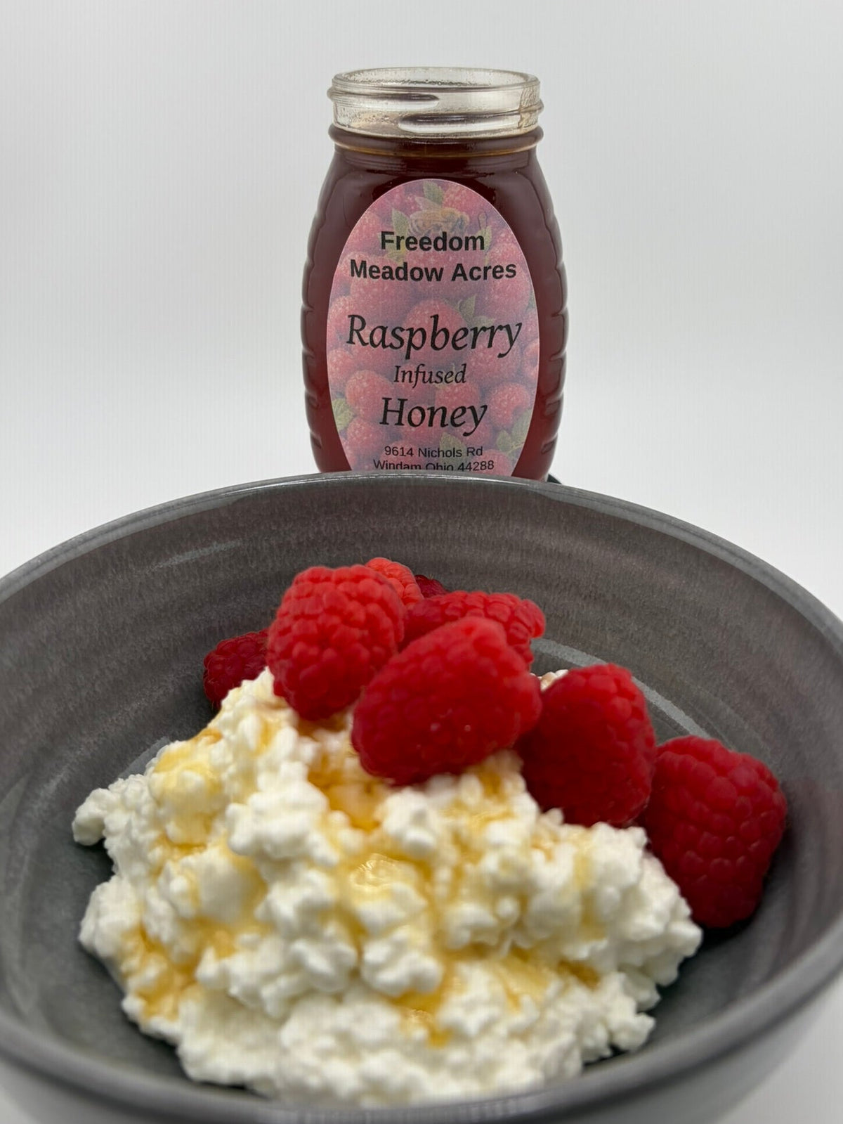 A jar of Freedom Meadow Acres Raspberry Infused Honey stands behind a dark gray bowl filled with white cottage cheese and topped with fresh red raspberries.