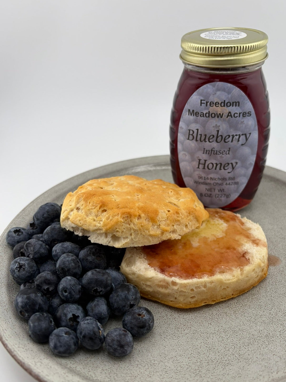 A jar of Freedom Meadow Acres Blueberry Infused Honey stands behind a plate with two golden-brown biscuits topped with blueberry infused honey and a side of fresh blueberries.
