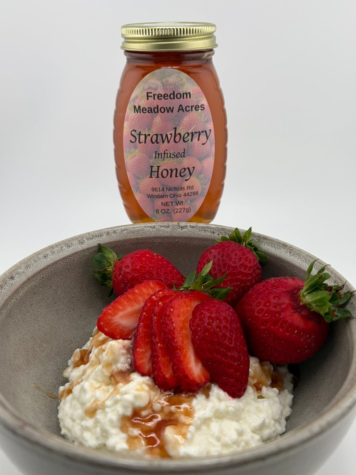 A jar of Freedom Meadow Acres Strawberry Infused Honey stands behind a gray ceramic bowl filled with white cottage cheese, sliced strawberries, and drizzled honey on a white background.