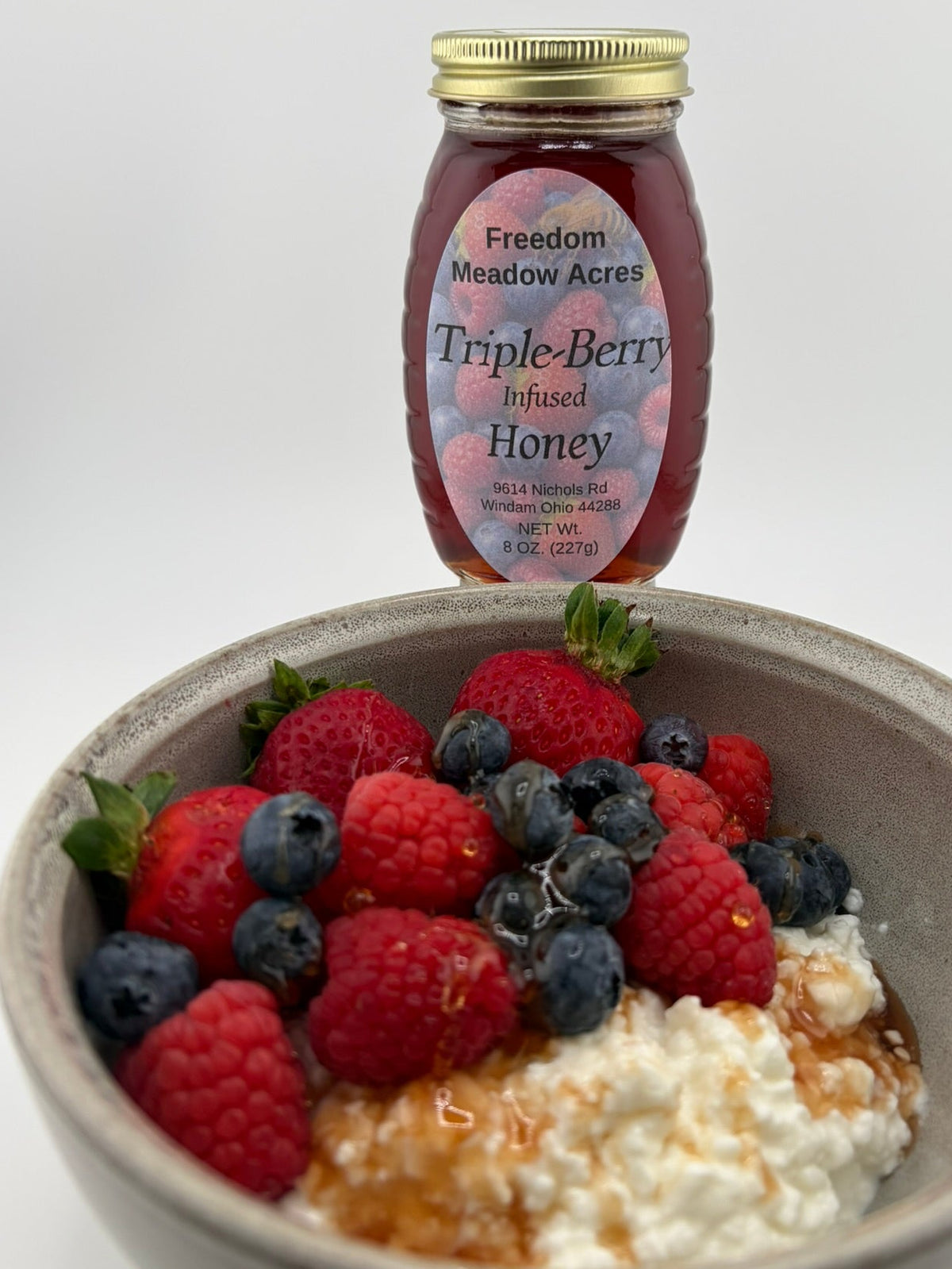 A jar of Freedom Meadow Acres Triple-Berry Infused Honey with a bowl filled with cottage cheese, strawberries, blueberries, and raspberries.
