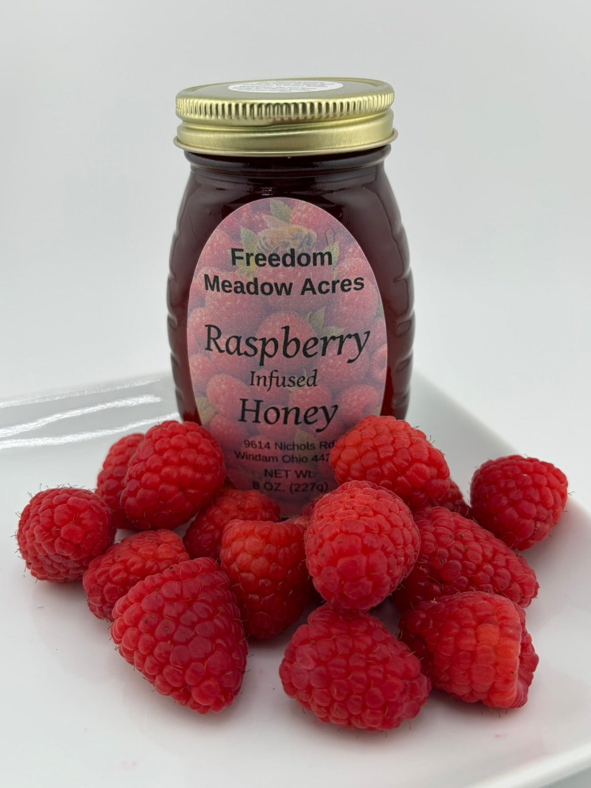 A jar of Freedom Meadow Acres Raspberry Infused Honey sits on a white plate surrounded by fresh red raspberries.