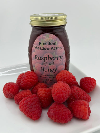 A jar of Freedom Meadow Acres Raspberry Infused Honey sits on a white plate surrounded by fresh red raspberries.