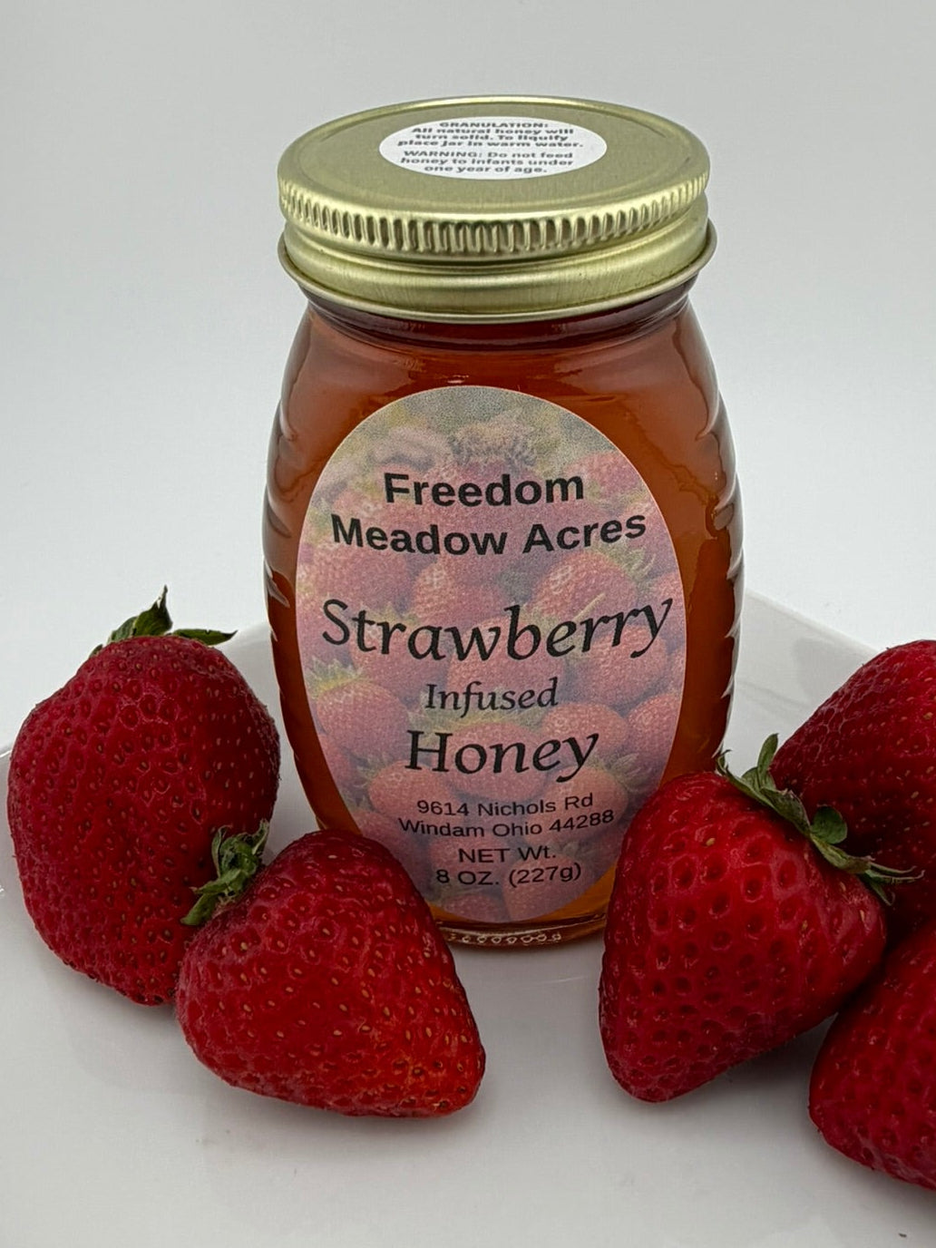 A glass jar of Freedom Meadow Acres Strawberry Infused Honey with a gold lid, displayed on a white tray with four fresh red strawberries around it against a light gray background.