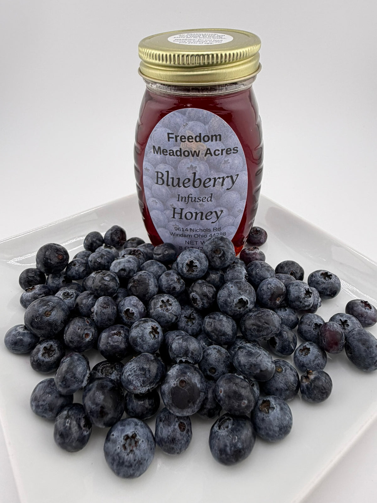 A glass jar of Blueberry Infused Honey by Freedom Meadow Acres sits on a plate piled with fresh blueberries.